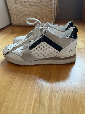 Vita sneakers från Michael Kors i storlek 38.5 - Snygga vita sneakers från Michael Kors  med svarta detaljer och perforerat mönster på sidorna. Skorna har grå mockadetaljer och en chunky sula som ger en sportig vibe. Perfekta för dig som gillar stilrena och trendiga sneakers. Storlek 38.5