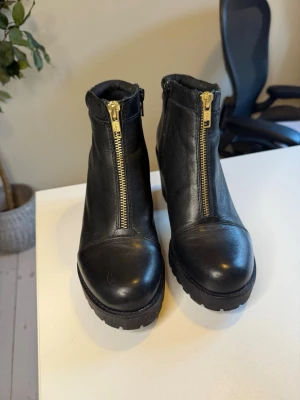 Svarta stövletter med gulddragkedja i läder Vagabond - Snygga svarta stövletter i skinn med chunky klack och grov sula. Dragkedja i guld framtill ger en cool och modern look. Skorna har rundad tå och vadderad kant upptill för extra komfort. Perfekta för dig som vill ha en edgy stil. Oanvända. Har några knappt märkbara småmärken efter att ha legat nedpackade, annars inget att anmärka på. Normala i storleken. Klackhöjd 8 cm men har en platå på 2 cm, så blir som en klack på 6 cm. Omhändertagna med lädervård.