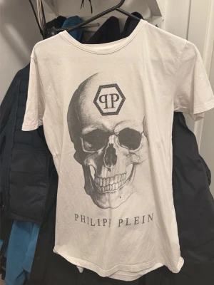Vit Philipp Plein t-shirt med dödskalle - Vit t-shirt från Philipp Plein med stort tryck av en dödskalle och PP-logga på bröstet. Klassisk rund halsringning och korta ärmar. Perfekt för dig som gillar edgy streetwear och vill sticka ut. Passar S/M/L