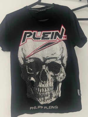 Svart t-shirt med dödskalle från Philipp Plein  ”Pris kan diskuteras”  - Cool svart t-shirt från Philipp Plein med stor glittrig dödskalle framtill och logga i vitt och rött. T-shirten har korta ärmar och rund hals. Perfekt för dig som gillar edgy streetstyle och vill sticka ut.
