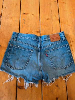 Levi's blå jeansshorts med fransar - Säljer ett par klassiska blå jeansshorts från Levi's med hög midja och råa, fransiga benslut. Shortsen har fem fickor, knappgylf och den ikoniska läderpatchen bak. Perfekta för sommardagar och en avslappnad stil.Storleken står inte i shortsen men passar perfekt på mig som är en S!