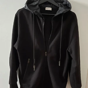 Moncler cardigan  - Svart hoodiejacka från Moncler med dragkedja framtill och två fickor med dragkedja. Jackan har huva med snörning och Moncler-logga på ärmen. Perfekt för en stilren och avslappnad look och nu till våren e den parfekt. Självklart äkta 