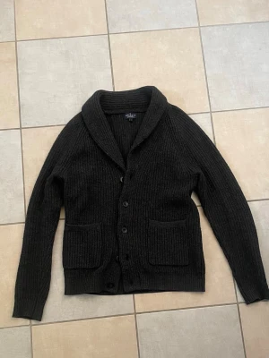 Hackett kofta  - Mysig svart stickad kofta från hackett med sjalkrage och stora framfickor. Koftan har ribbstickat mönster och knäpps med fem knappar framtill. Perfekt för lager-på-lager och chill dagar.