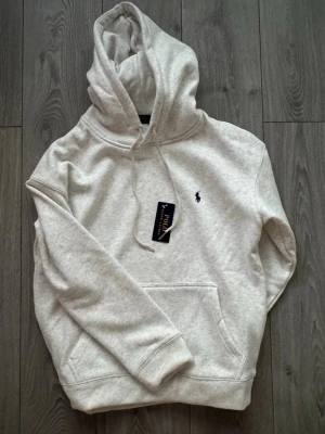 Polo Ralph Lauren helt ny  - Snygg ljusgrå hoodie från Polo Ralph Lauren med klassisk huva och känguruficka. Broderad logga i mörkblått på bröstet. Tillverkad i mjuk bomullsblandning som känns riktigt skön mot huden. Perfekt för dig som gillar stilrena och tidlösa plagg.