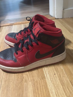 Air Jordan 1 Mid röd/svart - Säljer ett par Air Jordan 1 Mid i klassisk röd och svart färgkombination med svarta skosnören och vit sula. Skorna har ikoniska Jordan-loggor på plösen och sidan. Tillverkade i läder med perforerad tåbox för extra ventilation.