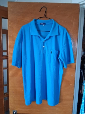 Blå piké från Polo Ralph Lauren - Klassisk ljusblå pikétröja från Polo Ralph Lauren med kort ärm, bröstficka och broderad logga. Skön bomullskvalitet och avslappnad passform. Perfekt för en chill och stilren look.