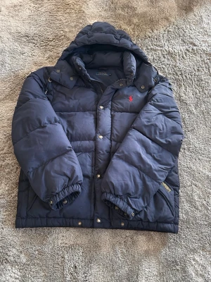 Mörkblå jacka Polo Ralph Lauren - Snygg mörkblå pufferjacka från Polo Ralph Lauren med huva och klassisk röd logga på bröstet. Jackan har dragkedja och tryckknappar framtill, elastiska muddar och är perfekt för kalla vinterdagar. Tjockt  vadderad för extra värme och stil. KOM MED BUD