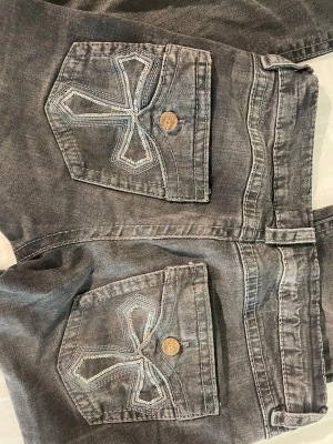 Grå bootcut jeans med broderade fickor - Snygga grå bootcut jeans med unika broderade bakfickor och guldiga knappar. Jeansen har en klassisk femficksmodell och lätt tvättad look. Perfekta för dig som gillar streetstyle och vill sticka ut med detaljer på fickorna. Ena knappen på bakfickan är lite lös, men sitter fortfarande på stabilt! Skriv vid frågor, pris går att diskutera!💕💕 