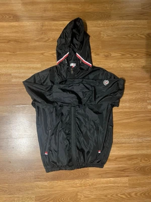 Svart Moncler vindjacka med huva - Svart vindjacka från Moncler med huva och dragkedja framtill. Jackan har röda och vita detaljer på huvan och små logotyper på bröstet och vid fickorna. Tillverkad i ett lätt, glansigt material som står emot vind. Perfekt för en sportig och stilren look.