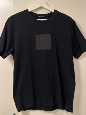 Svart t-shirt från C.P. Company - Svart t-shirt från C.P. Company med en stilren patch på bröstet där loggan är präglad. Klassisk rund halsringning och korta ärmar. Tillverkad i mjuk bomull för en bekväm känsla och enkel stil.
