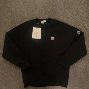 Svart sweatshirt från Moncler - Svart sweatshirt från Moncler med rund halsringning och klassisk Moncler-logga på bröstet samt ärmen. Tröjan har en enkel och stilren design med ribbade muddar vid ärmslut och nederkant. Perfekt för en clean och trendig look.