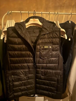 Svart dunväst från Barbour International - Snygg svart dunväst från Barbour International med huva, dragkedja och flera praktiska fickor framtill. Västen har quiltad design och en bröstficka med tryckknappar och logotyp. Perfekt för lager-på-lager och streetstyle.