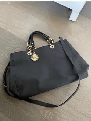 Svart Michael Kors handväska - Snygg svart handväska från Michael Kors med guldfärgade detaljer och kedjor. Väskan har både handtag och en längre axelrem, samt en rund MK-logga i metall. Tillverkad i slätt läder och har en klassisk, rymlig form. Kan finnas små spår av defekter men syns knappt😊Äkta