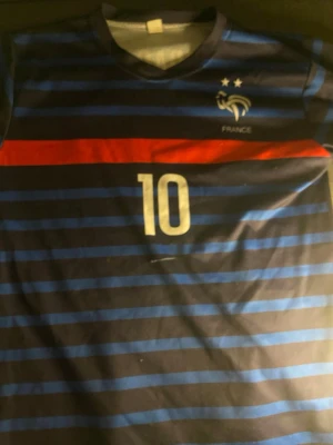 Frankrike fotbollströja Mbappé #10 - Säljer en blå och svart randig fotbollströja från Nike med Frankrikes landslagsmärke, två stjärnor och röd detalj. Nummer 10 och Mbappé tryckt på ryggen. Perfekt för dig som älskar fotboll och vill stötta Les Bleus.