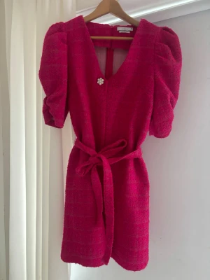 Rosa kortklänning med puffärm & bälte - Supersöt rosa kortklänning  med puffärmar, v-ringning och dekorativ blomknapp. Klänningen har ett matchande bälte i midjan och är tillverkad i ett strukturerat tyg som ger en exklusiv känsla. Perfekt för dig som vill sticka ut! Använd endast en gång. Storleken passar S/M