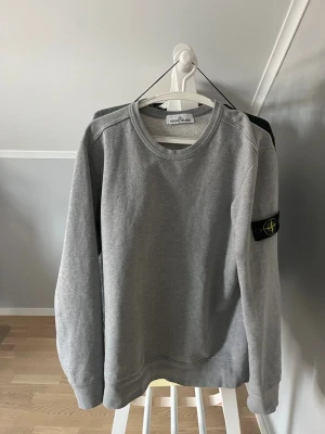 Grå sweatshirt från Stone Island - Säljer en stilren grå sweatshirt från Stone Island med klassisk rund hals och den ikoniska svarta logotypen med kompass på vänster ärm. Tröjan har ribbade muddar vid ärmslut och nederkant. Perfekt för en avslappnad och trendig look.
