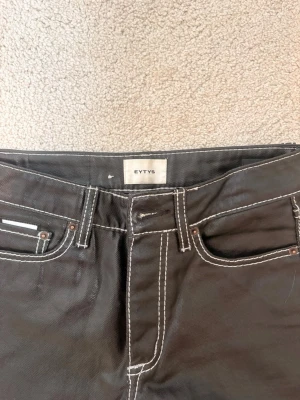 Svarta jeans från Eytys med kontrastsömmar - Säljer ett par svarta jeans från Eytys med raka ben och vita kontrastsömmar. Jeansen har klassisk femficksdesign och unika broderade detaljer på bakfickorna. Materialet är denim med en lätt glansig finish. Perfekta för en trendig och stilren look.