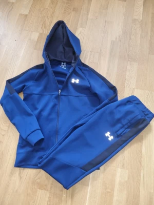Under armarna dress - Mysigt blått loungewear-set från Under Armour med matchande hoodie och byxor. Setet har dragkedja, huva och svarta detaljer längs ärmar och ben. Både tröjan och byxorna har logga framtill. Perfekt för chill dagar hemma.
