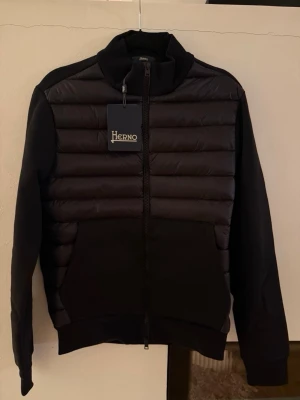 Svart pufferjacka från Herno - Snygg svart pufferjacka från Herno med quiltad framsida och släta ärmar. Jackan har hög krage, dragkedja framtill och stora fickor. Materialmix med vadderad front och mjuka partier på ärmar och rygg. Perfekt för kalla dagar.