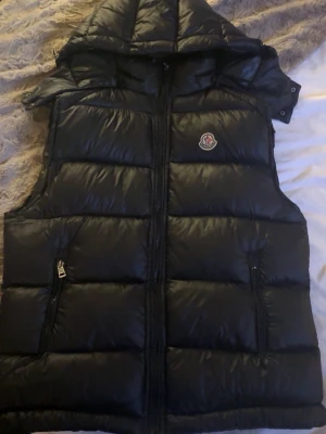 Dunväst - Moncler - Svart - Snygg svart dunväst från Moncler med avtagbar huva och dragkedja framtill. Västen har två fickor med dragkedja och Moncler-logga på bröstet. Perfekt för lager-på-lager och håller dig varm med sin vadderade design. Den har för det mesta bara hängt sen jag köpte den, den är i acceptabelt skick och har inga hål eller jättetydliga tecken på användning.