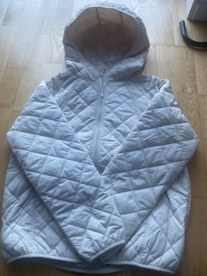 Créme färgad  quiltad pufferjacka med huva - Säljer en Créme färgad pufferjacka med quiltat mönster och dragkedja framtill. Jackan har en stor huva och är tillverkad i ett glansigt, vadderat material som håller värmen. Perfekt för kalla vinterdagar och riktigt snygg till streetwear-stilen.