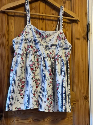 Blommig somrig topp med breda axelband - Söt topp med breda axelband i vitt tyg med blommigt mönster i rosa, rött och blått. Toppen har en luftig passform och vertikala ränder med små blommor. Perfekt för varma dagar och ger en romantisk vibe.