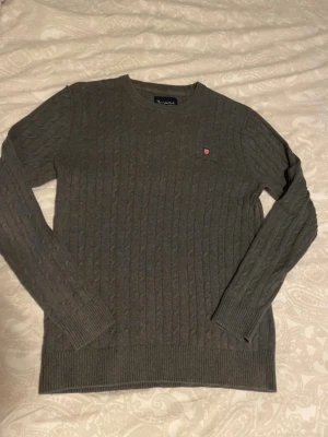 Mörkgrå kabelstickad tröja Bondelid - Snygg mörkgrå kabelstickad tröja från Bondelid med rund halsringning och diskret röd logga på bröstet. Tröjan har långa ärmar och ribbade muddar vid ärmslut och nederkant. Perfekt för dig som gillar klassisk och stilren design.