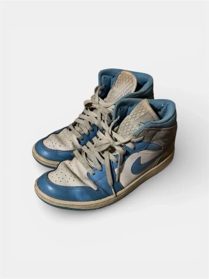 Nike Jordan 1 university blue grey mid  - Säljer ett par Nike Jordan 1 sneakers i blått, grått och vitt. Klassisk high-top modell med ikonisk swoosh på sidan och Air Jordan-logga vid ankeln. Ovandelen är i läder och textil, med perforerad tåbox och vadderad krage för extra komfort. Storlek: 42