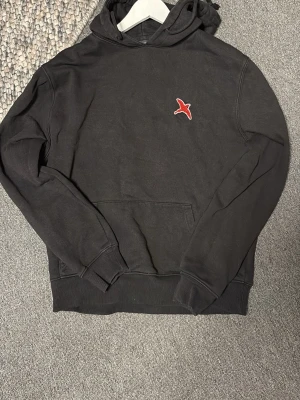 Svart hoodie från Axel Arigato - Snygg svart hoodie från Axel Arigato med röd broderad fågel på bröstet. Klassisk känguruficka framtill, ribbade muddar och huva. Perfekt för en avslappnad och trendig look. Tillverkad i mjukt bomullsmaterial. Lite tvättblekt men fortfarande fin