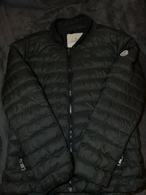 Grön dunjacka från Moncler - Snygg grön dunjacka från Moncler med quiltad design och två dragkedjeförsedda fickor framtill. Jackan har hög krage och klassisk Moncler-logga på ärmen. Storlek XL men sitter som M 