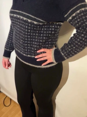 Mörkblå stickad tröja med mönster - Mysig mörkblå stickad tröja med grått och vitt mönster över bröst, ärmar och midja. Tröjan har hög krage och ribbstickade muddar vid ärmslut och nederkant. Perfekt för kalla dagar och har en klassisk nordisk känsla.