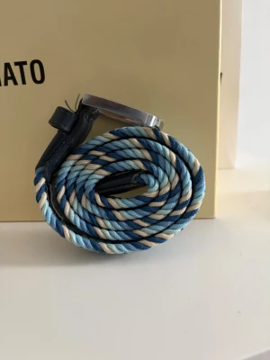 Randigt flätat bälte i blå toner - Snyggt flätat bälte från Velasca med randigt mönster i ljusblått, mörkblått och beige. Bältet har detaljer i svart skinn och ett klassiskt metallspänne. Perfekt accessoar för att lyfta din outfit med lite färg. För mer info kontakta gärna! ABS! Pris går att diskutera! 