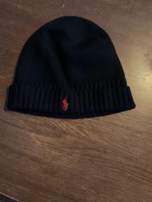 Svart mössa från Polo Ralph Lauren - Snygg svart mössa från Polo Ralph Lauren med uppvikt kant och klassisk röd logga broderad framtill. Tillverkad i mjukt stickat material som håller dig varm och stilren under kalla dagar.