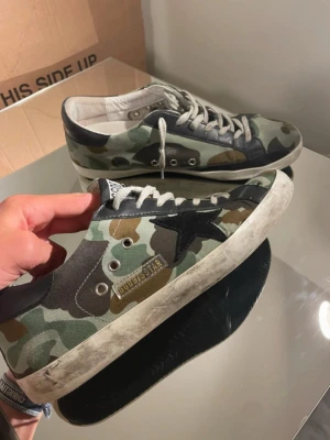 Golden Goose - Snygga låga sneakers från Golden Goose med camo-mönster i grönt, brunt och svart. Skorna har GGDB-logga på plösen, svart stjärndetalj på sidan och slitna detaljer på sulan för en cool, streetig vibe. Perfekta för dig som gillar unik stil och italiensk design.
