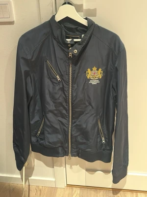 Morris marinblå bomberjacka - Snygg marinblå bomberjacka från Morris med broderad logga och vapensköld på bröstet. Jackan har snedställda dragkedjefickor, ribbade muddar och hög krage med tryckknapp. Tillverkad i glansigt syntetmaterial som ger en sportig look.