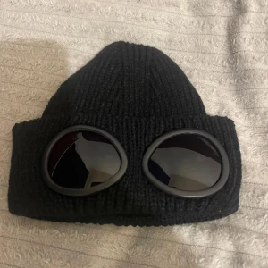 Svart C.P. Company goggle-mössa - Säljer en svart stickad mössa från C.P. Company med de ikoniska inbyggda goggles framtill. Mössan är ribbstickad och har en cool, urban vibe som sticker ut. Perfekt accessoar för dig som vill ha något unikt och trendigt.