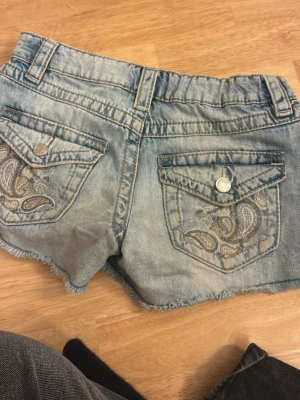 Ljusa jeansshorts med broderade fickor - Snygga ljusblå jeansshorts med slitna detaljer och råa kanter. Baksidan har två fickor med paisleybroderi och knappstängning. Klassisk femficksmodell med bälteshällor och dragkedja framtill. Perfekta för sommardagar!