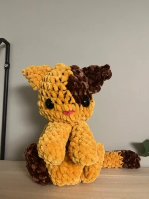 Gul och brun virkad kattmjukis - Supergullig handvirkad katt i mjuk plysch med stora svarta ögon, gul och brun färg samt rosa nos. Perfekt som prydnad eller gosedjur. Katten har lång svans och stora öron.