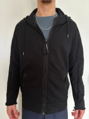 Svart hoodie med dragkedja och goggles - Svart hoodie med hel dragkedja, två fickor med dragkedja och justerbar huva med inbyggda goggles baktill. Snygg streetstil och cool detalj med glasögon på huvan. Perfekt för dig som gillar unika plagg.