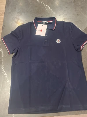 Marinblå pikétröja från Moncler - Snygg marinblå pikétröja från Moncler med klassisk krage och knappar framtill. Tröjan har Moncler-logga på bröstet och röda samt vita detaljer på krage och ärmslut. Perfekt för en stilren look.