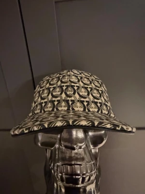 Beige keps med svart mönster från Dior - Säljer en beige keps från Dior med svart grafiskt mönster och böjd skärm. Kepsen har klassisk form och är tillverkad i ett tygmaterial med tydligt logomönster. Perfekt för dig som vill sticka ut med en exklusiv accessoar.(ÄKTA) träffas oxå i Sthlm 