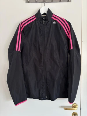 Svart vindjacka med rosa ränder Adidas - Snygg svart vindjacka från Adidas med ikoniska rosa ränder längs axlar och ärmar. Jackan har hög krage, dragkedja framtill och är tillverkad i lätt, vindtätt material.