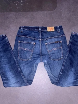 Blå Nudie Jeans med klassisk passform - Säljer ett par blå jeans från Nudie Jeans med klassisk femficksdesign och snygga detaljer på bakfickorna. Jeansen har en mörkblå tvätt och är tillverkade i denim. Perfekt för dig som gillar stilrena och tidlösa jeans. 33/32