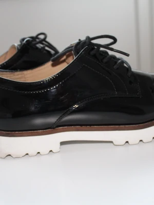 Svarta lackloafers med vit sula - Snygga loafers i svart lack med snörning och chunky vit sula. Skorna har en glansig finish, beige insida och en kontrasterande brun detalj mellan sula och ovandel. Perfekt för dig som gillar en modern och edgy stil.