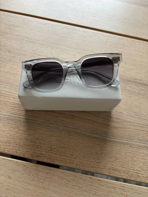 Transparenta solglasögon från Chimi - Säljer ett par stilrena solglasögon från Chimi med transparenta bågar och mörka linser. Glasögonen har en fyrkantig form och kommer med ett vitt hårt fodral. Perfekt för dig som vill ha en modern och clean look.