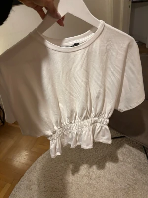 Vit croppad topp med rynkdetalj - Säljer en vit croppad topp med korta ärmar och rynkad midjedetalj som ger en snygg volangkant. Perfekt till jeans eller kjol. Enkel och stilren design i mjukt material🩷