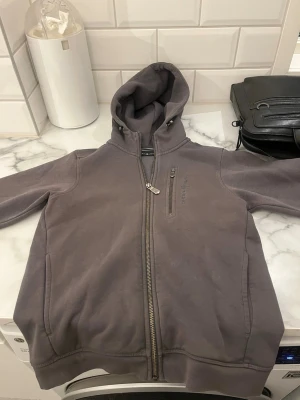 Mörkgrå hoodie med dragkedja - Säljer en mörkgrå hoodie från Peak Performance med hel dragkedja och huva. Tröjan har en bröstficka med dragkedja och ribbade muddar. Perfekt för chill dagar och enkel att matcha med jeans eller joggers.