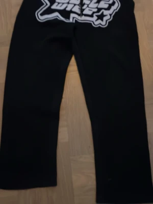 Svarta mjukisbyxor Wide World - Svarta mjukisbyxor med bred passform och stort vitt tryck framtill med texten 'Wide World' och stjärndetalj. Byxorna har raka ben och är tillverkade i mjukt material, perfekta för en avslappnad stil.