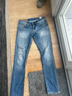 Blå slitna jeans från dondup jeans - Ett par blå jeans från dondup jeans med slitningar och ljusa tvättade partier. Klassisk femficksmodell med knappgylf och läderpatch med logga bak i midjan. Jeansen har raka ben och kontrastsömmar för en avslappnad look. Passar 30