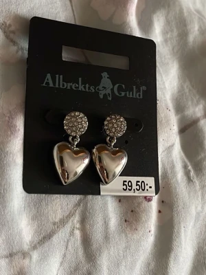 Silverfärgade hjärtörhängen Albrekts Guld - Snygga örhängen i silverfärg från Albrekts Guld med glittrande stenar på knoppen och hängande hjärtan. Perfekta för dig som vill ha något som sticker ut och ger lite extra bling till din stil.